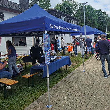 Blaues Duratent Faltzelt Hexa Compact mit Aufdruck „SV Adler Dellbrück 1920 e.V. Jugendabteilung“ bei einem Vereinsfest. Unter dem Faltpavillon bieten Tische mit Speisen und Getränken den Gästen Verpflegung. Ideal für Sportvereine und Outdoor-Veranstaltungen.